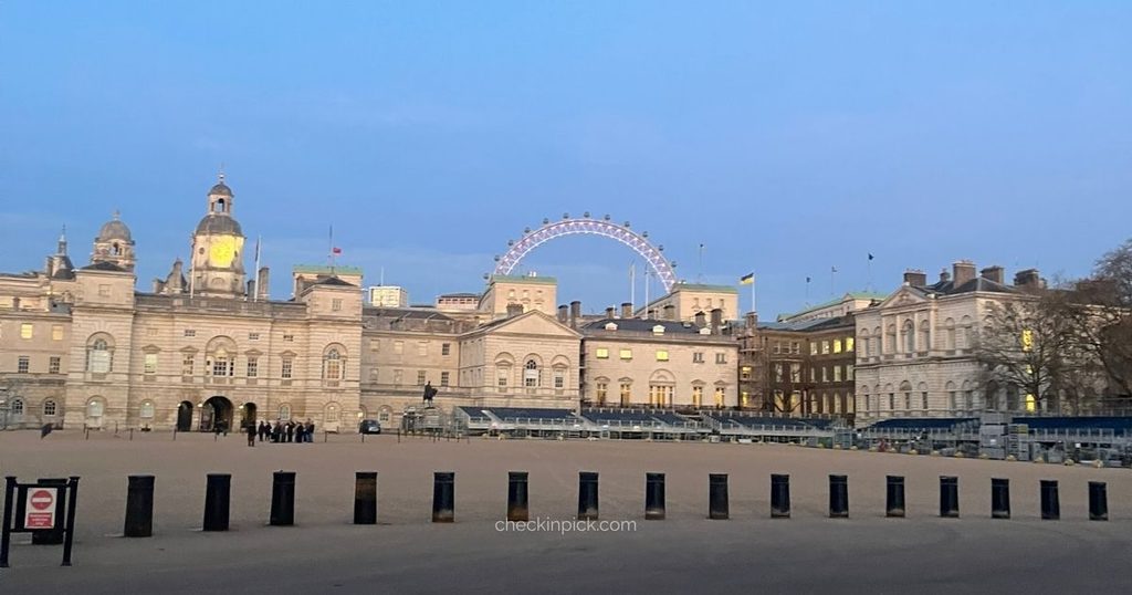 A wide-angle view of Horse Guards Parade in London at dusk, featuring historic white buildings and the London Eye glowing softly under a clear blue evening sky.